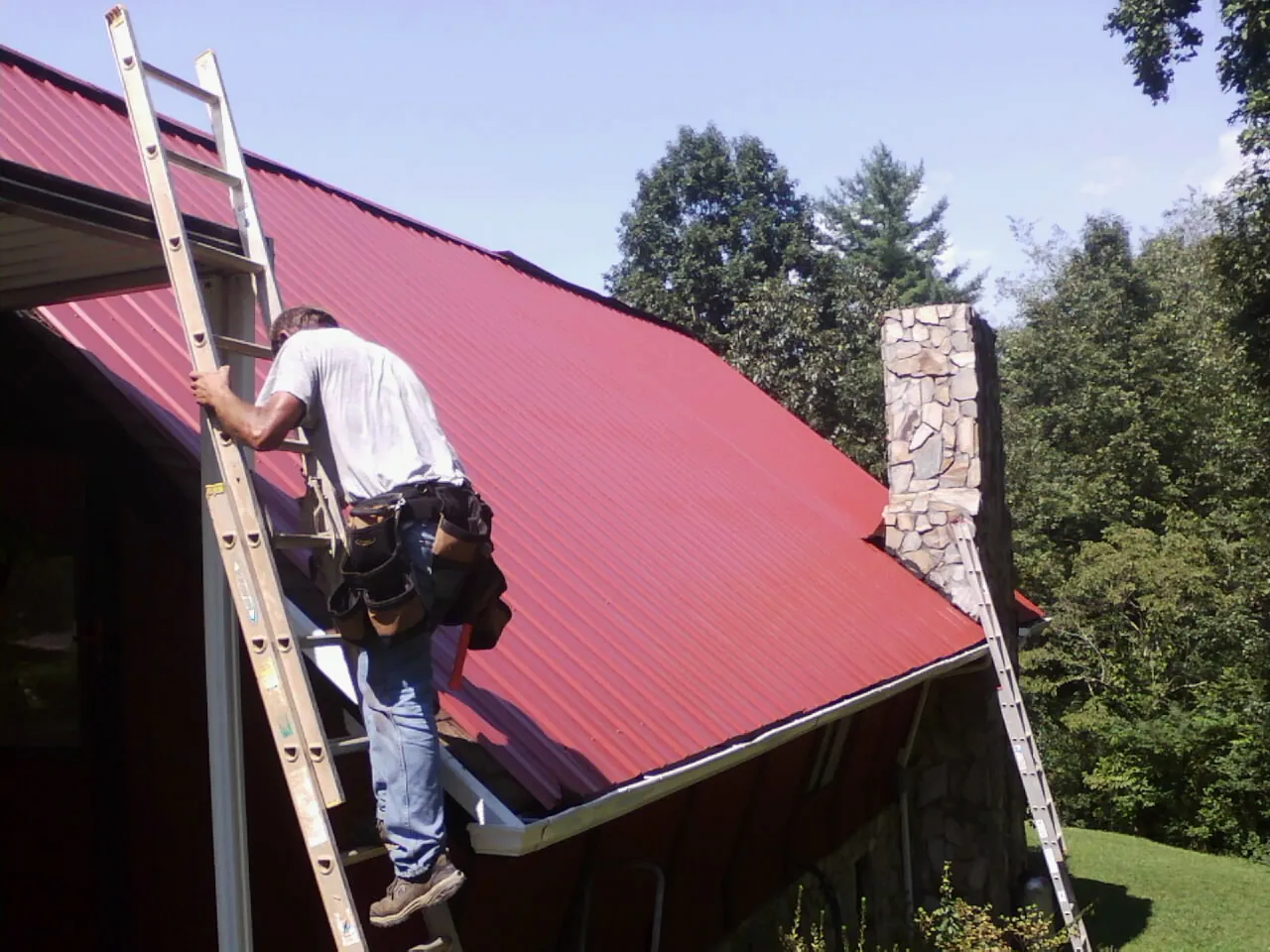 Roofer on a ladder beside a red metal roof and stone chimney — Soffit & Fascia Repair by Roofing Brothers in Boalsburg